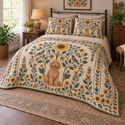 A curly goldendoodle sits beneath a tall folk sunflower, surrounded by mirrored leaves, smaller blooms and birds on warm cream on a quilt set with two matching shams, angled view.