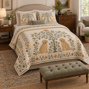 Two small goldendoodles sit on opposite sides of a flowering folk tree, framed by cream, sage and golden leaf motifs on a quilt set with two matching shams, angled view.