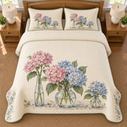 AI mockup, high overhead bedroom view of a hydrangea quilt with hydrangeas arranged in three glass vases and two matching printed pillow covers.